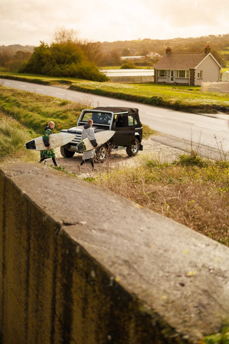 Colour photo of a person and a boy carrying surfboards on a road