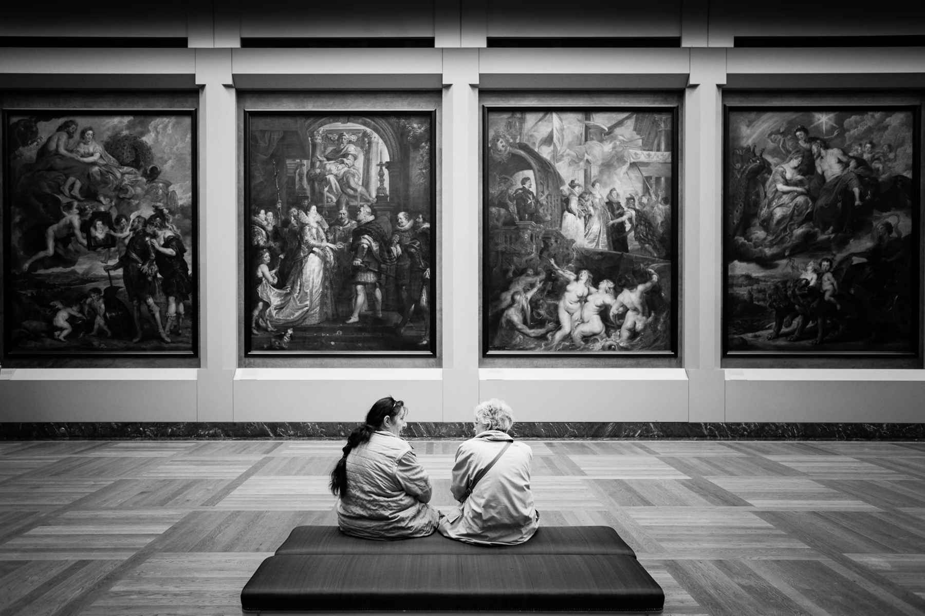 Black & white photo of a couple of people sitting on a bench looking at a wall of paintings