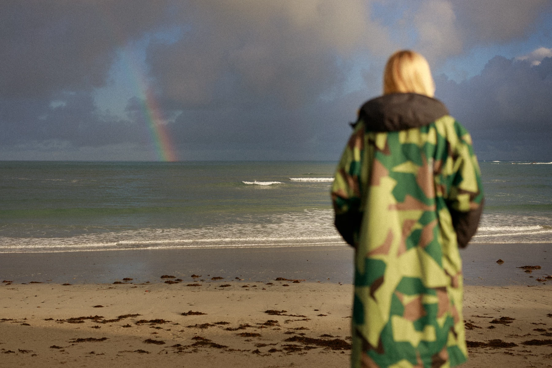 Colour photo of a person standing on a beach looking at the water with a rainbow in the sky